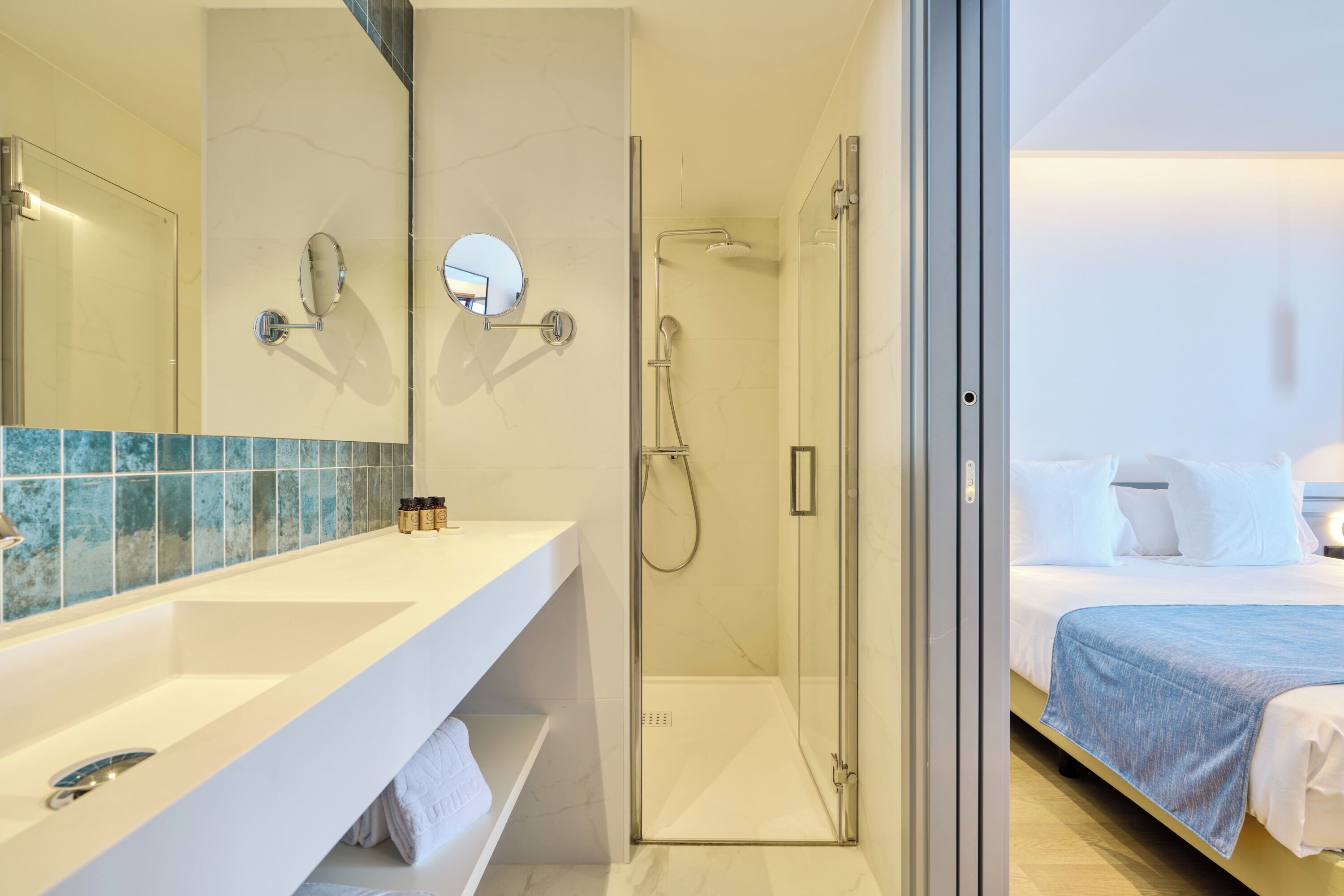 A modern bathroom with a glass shower enclosure, a white vanity with a mirror, and a bed with a blue blanket in the adjacent room.