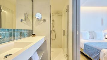A modern bathroom with a glass shower enclosure, a white vanity with a mirror, and a bed with a blue blanket in the adjacent room.