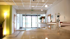 Un vestíbulo de hotel con suelo de baldosas, una recepción, plantas en macetas y una puerta de cristal que da al exterior.