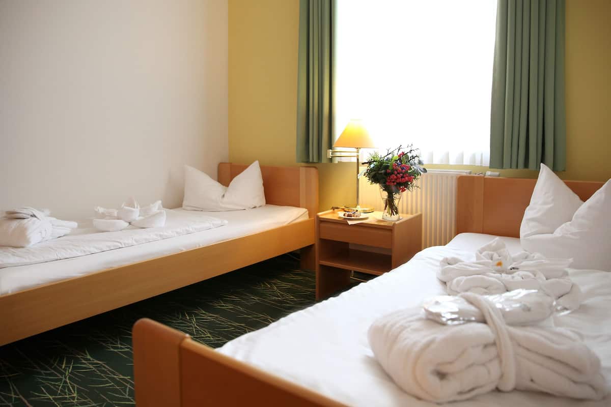 A hotel room with two beds, a nightstand with a lamp and flowers, and a window with curtains.