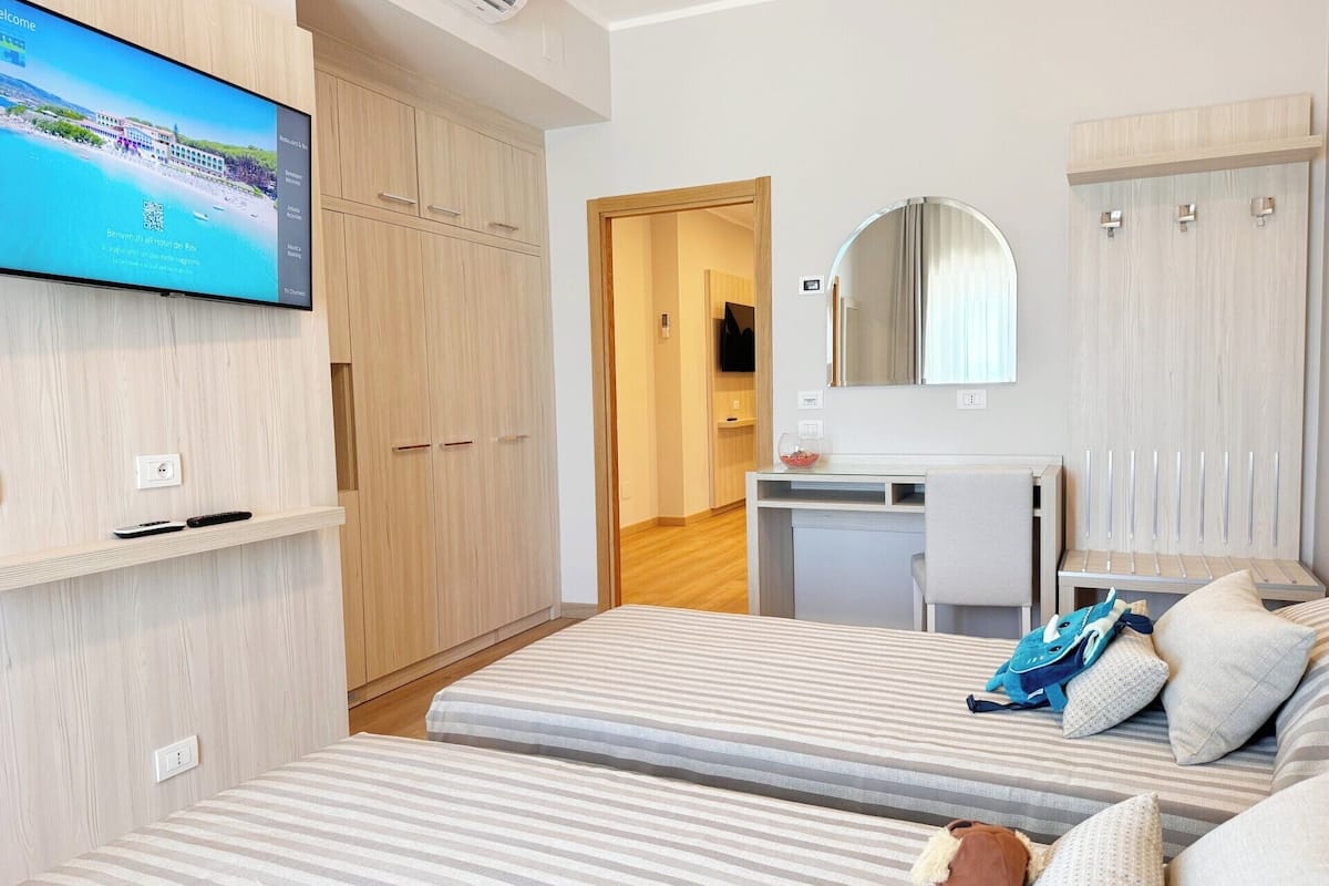 Ein Hotelzimmer mit Fernseher, der eine Strandansicht zeigt, einem Kleiderschrank, einem Schreibtisch mit Spiegel und zwei Betten mit gestreifter Bettwäsche.