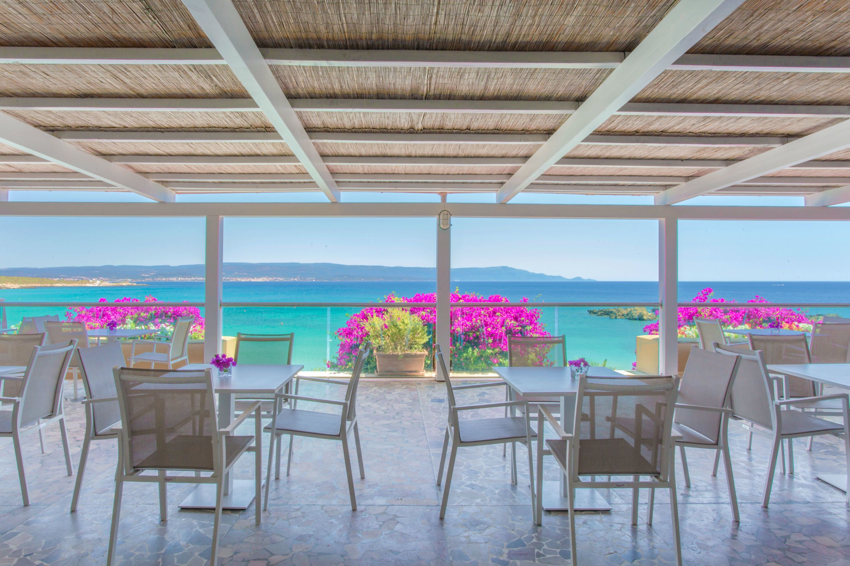 An outdoor terrace with tables and chairs, overlooking a coastal view with mountains and the sea.