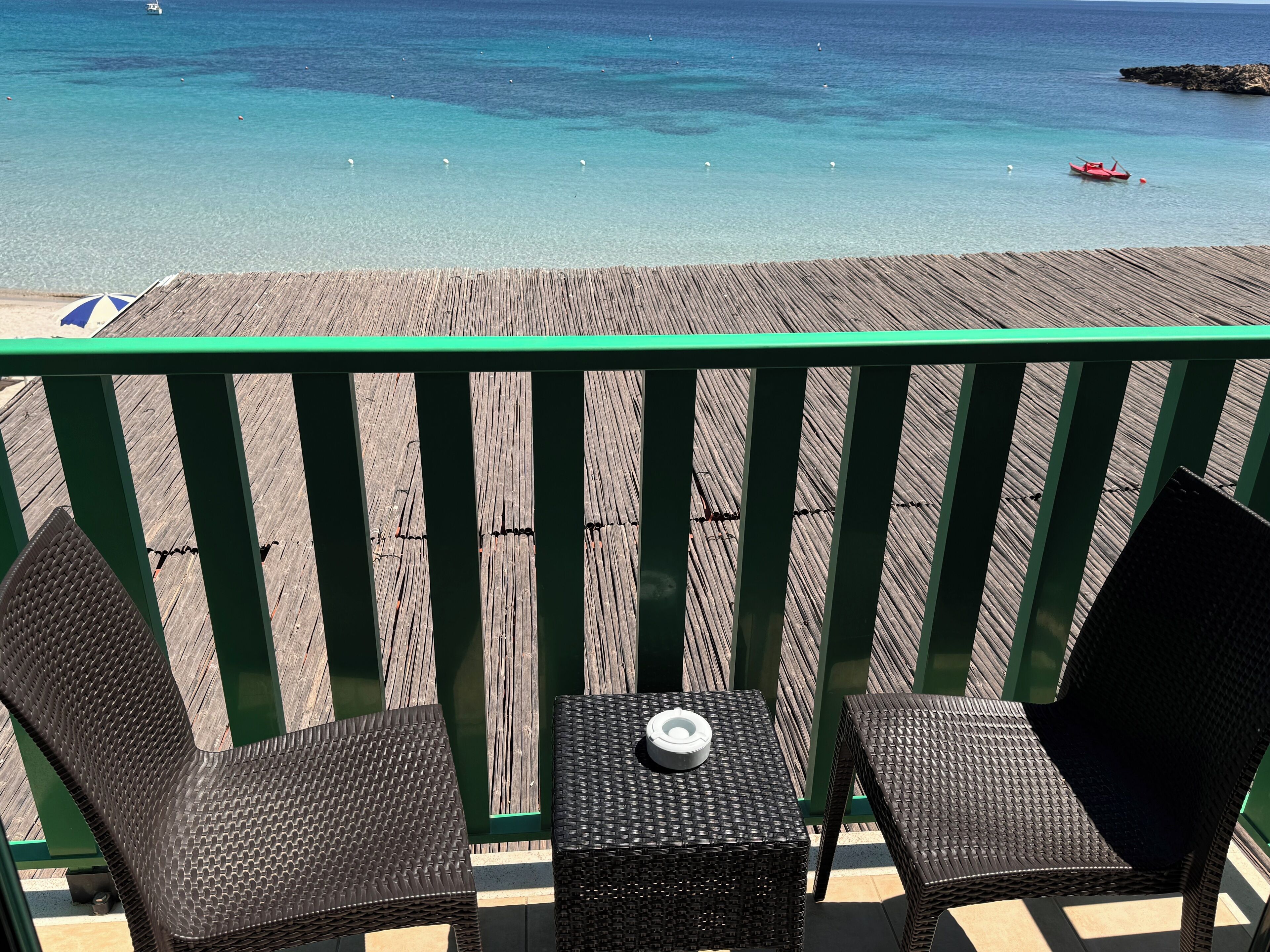 A wooden deck with two wicker chairs and a small table, overlooking a clear blue sea with a boat and rocky outcrop.