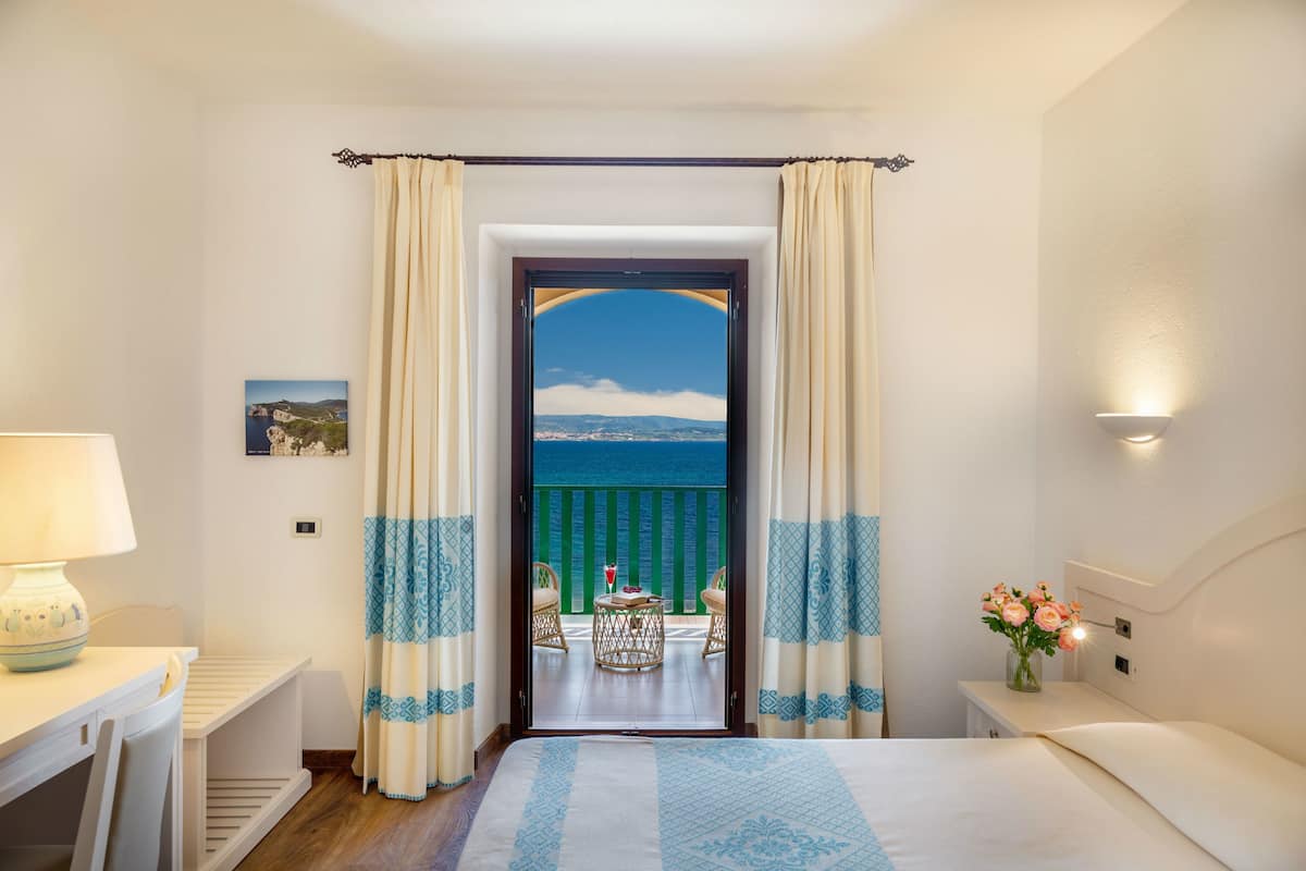 Ein Hotelzimmer mit Bett, Schreibtisch, Stuhl, einem kleinen Tisch mit einem Getränk und Meerblick durch eine Tür.