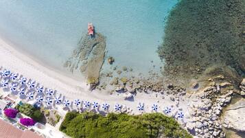 Vista aerea di una spiaggia con acque limpide, zone rocciose e una fila di ombrelloni blu e bianchi.