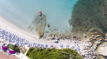 Vista aerea di una spiaggia con acque limpide, zone rocciose e una fila di ombrelloni blu e bianchi.