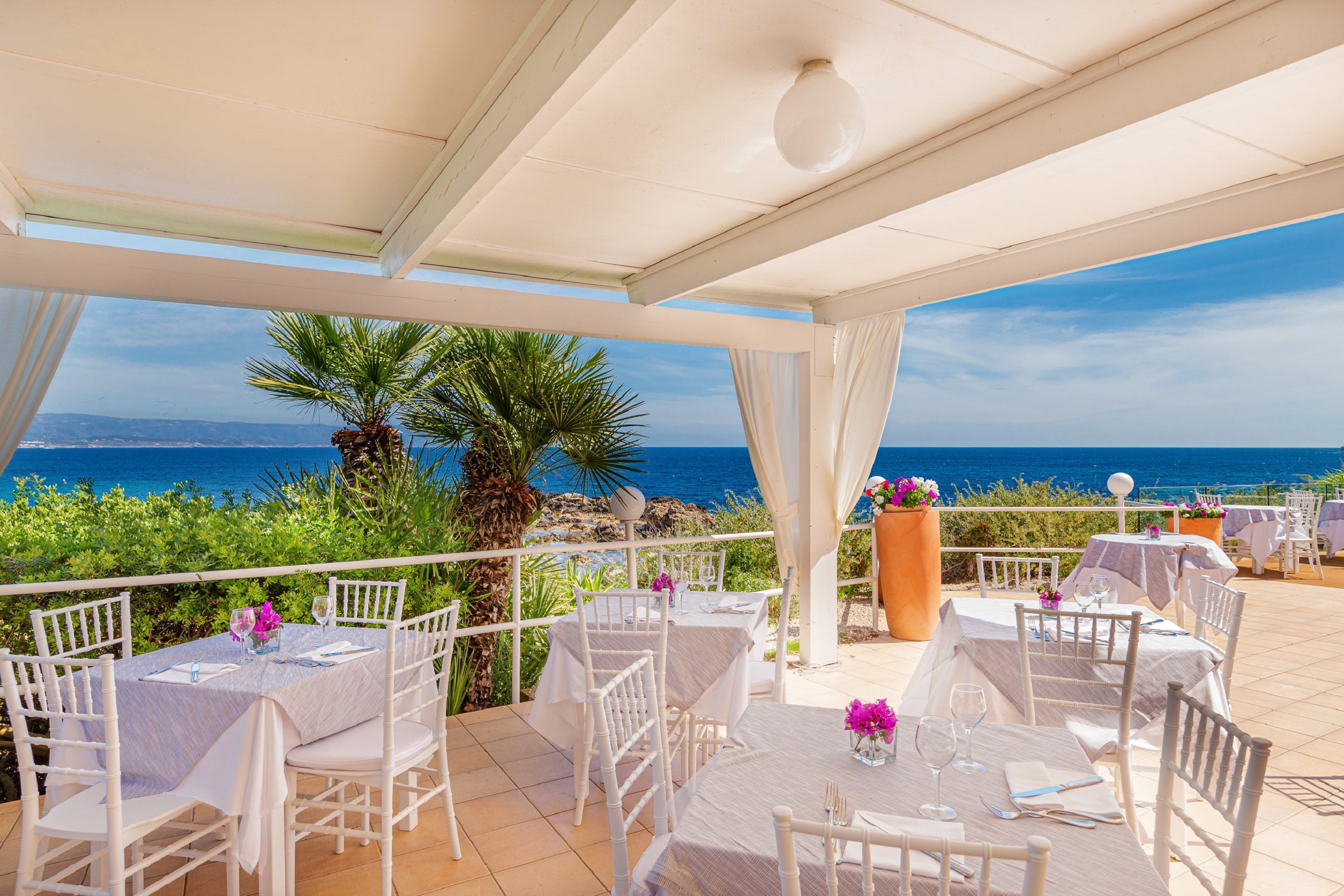 An outdoor dining area with white tables and chairs, overlooking a scenic ocean view.