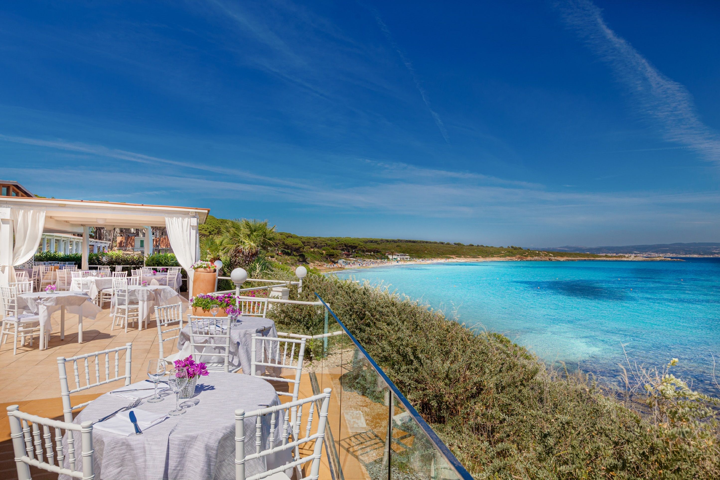 An outdoor dining area with white chairs and tables, overlooking a beach and the sea.