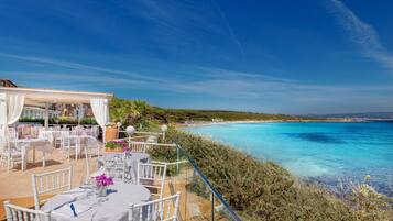 An outdoor dining area with white chairs and tables, overlooking a beach and the sea.