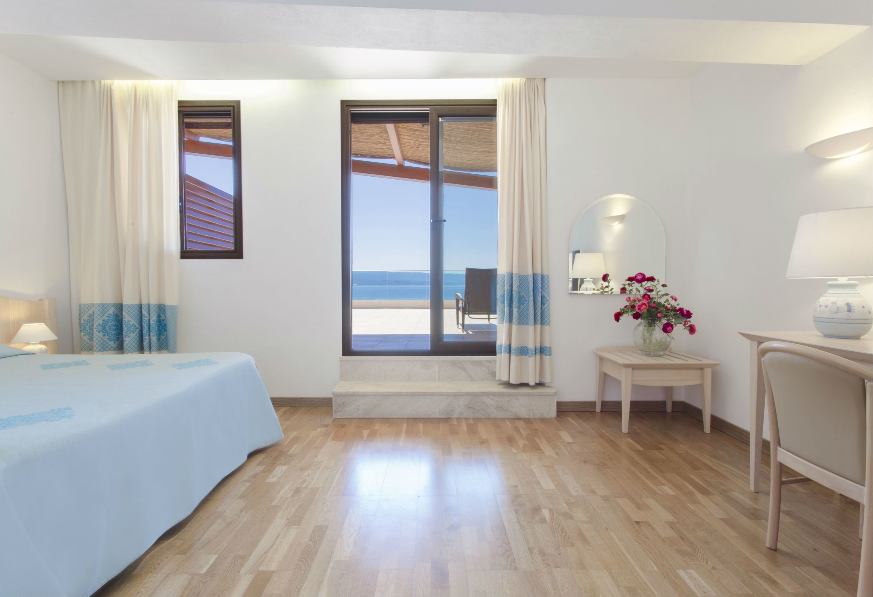 Une chambre d’hôtel avec un lit, une petite table avec une lampe, un miroir, un vase avec des fleurs et une vue sur la mer, visible à travers une porte coulissante.