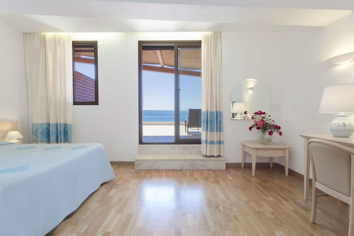 Une chambre d’hôtel avec un lit, une petite table avec une lampe, un miroir, un vase avec des fleurs et une vue sur la mer, visible à travers une porte coulissante.