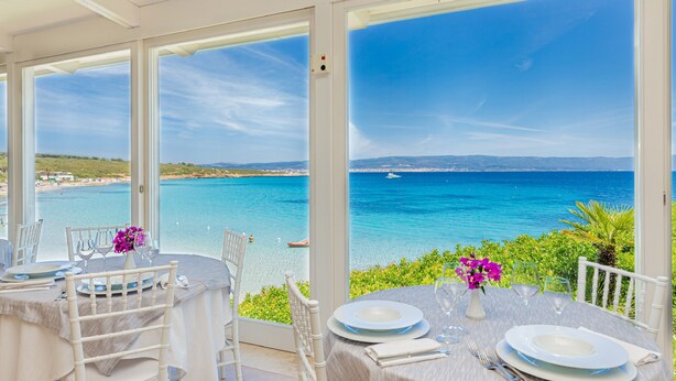Un área de comedor al aire libre con una mesa dispuesta para dos, con vistas a la costa y aguas azules cristalinas, y tierra a lo lejos.
