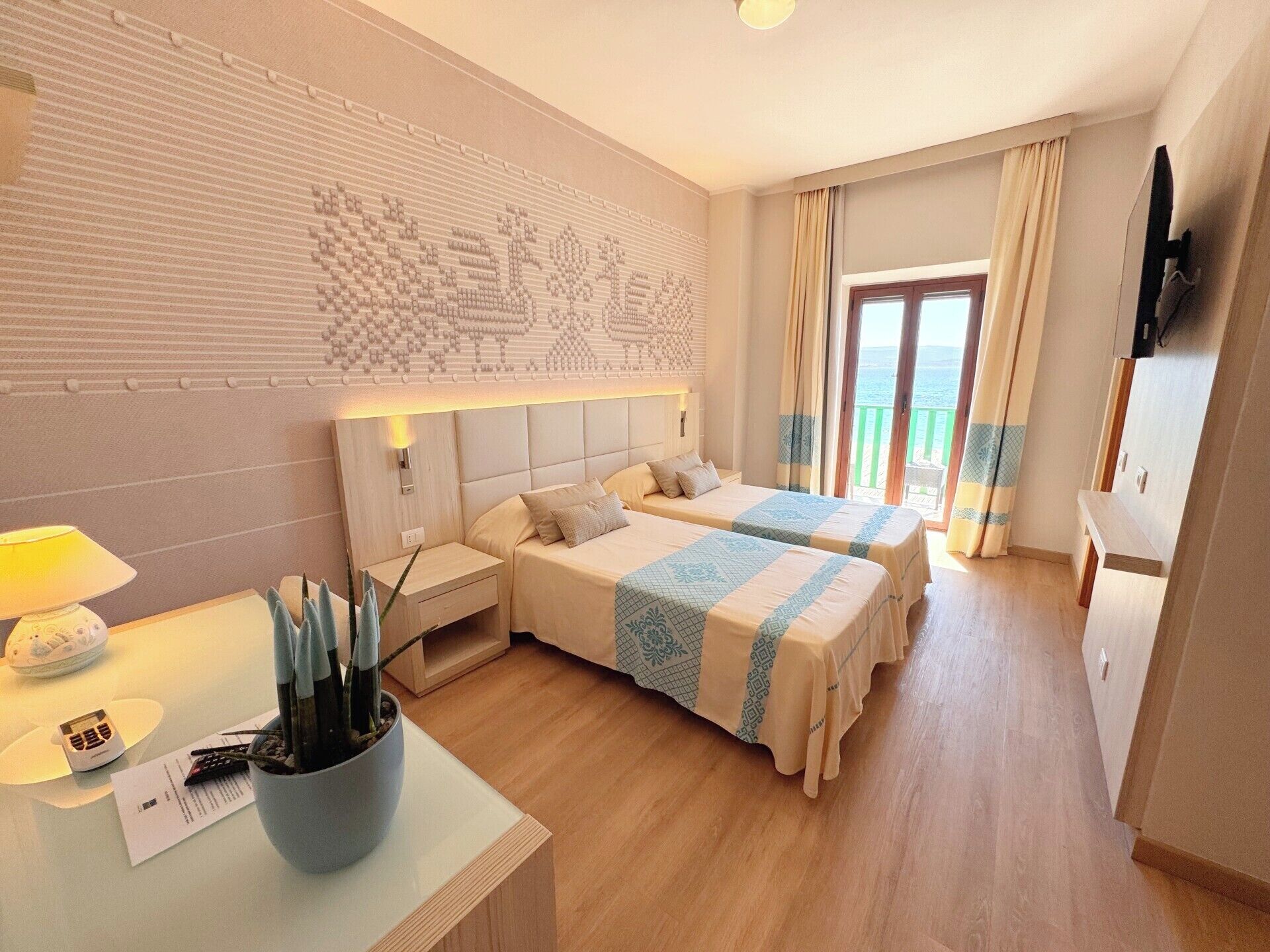 A hotel room with two beds, a large window with curtains, a flat-screen TV, and a small table with a lamp and a plant.