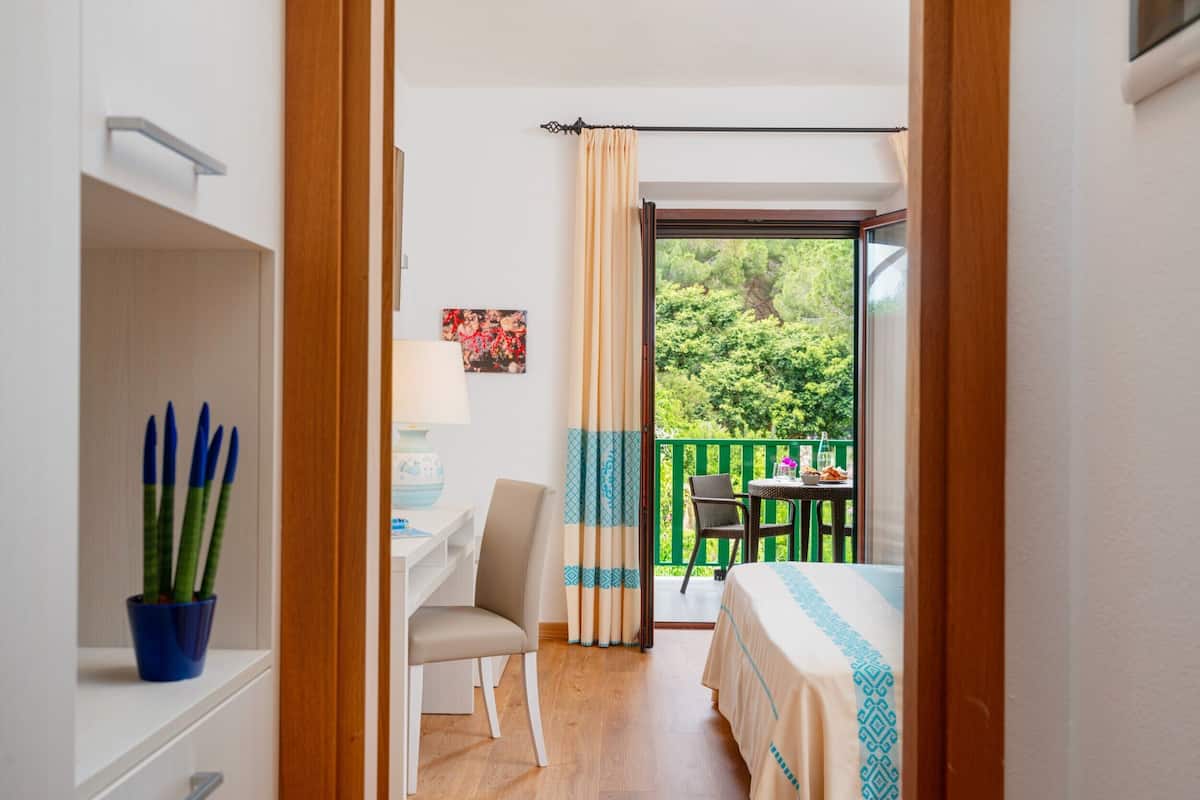 Ein Hotelzimmer mit Schreibtisch, Stuhl und Blick auf einen Balkon mit Grünpflanzen.