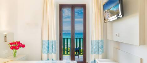 A bedroom with a bed, a TV, a window with curtains, and a view of the sea.