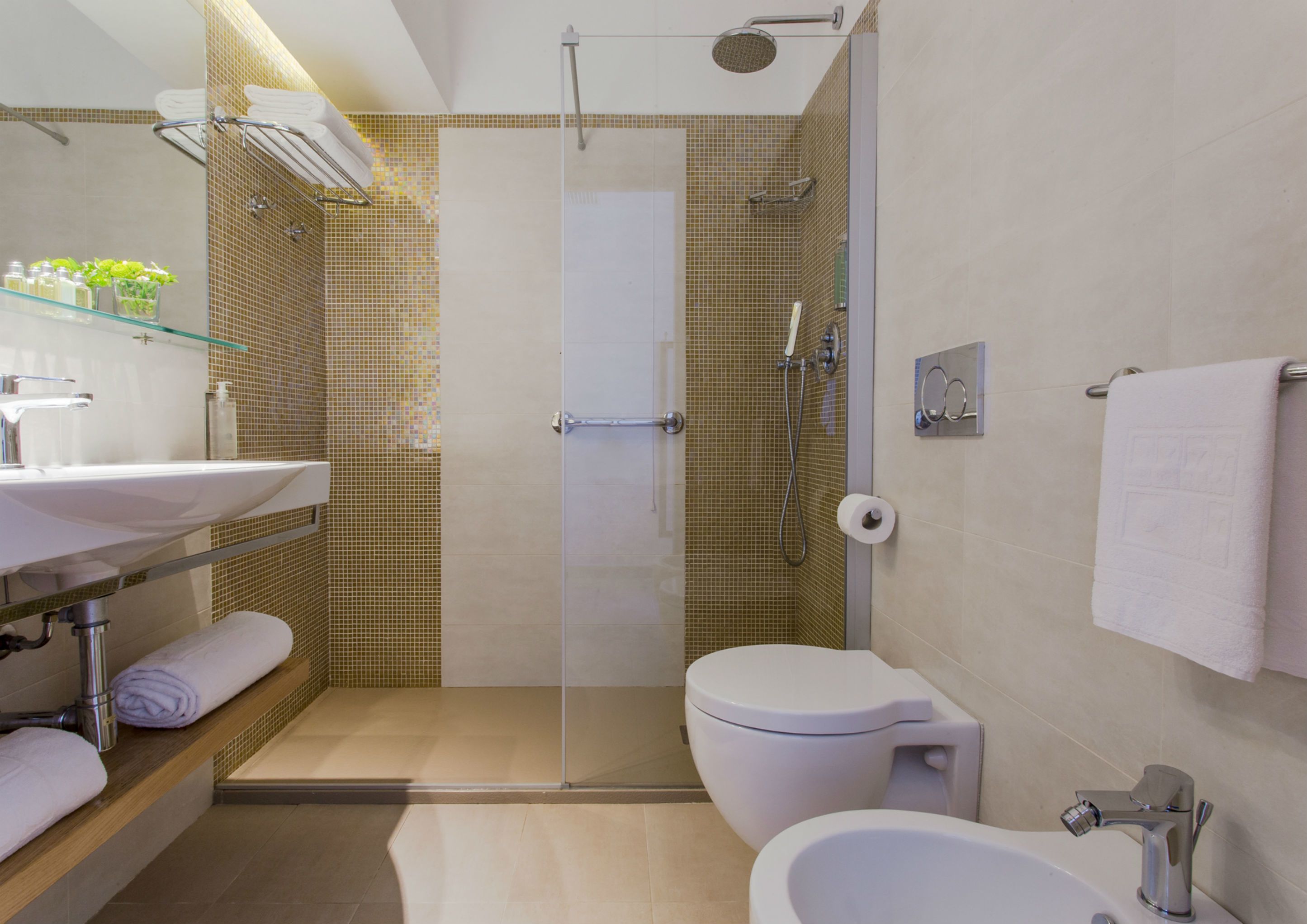 Un baño moderno con ducha de vidrio, inodoro, bidé y lavamanos con repisas de vidrio para artículos de aseo.