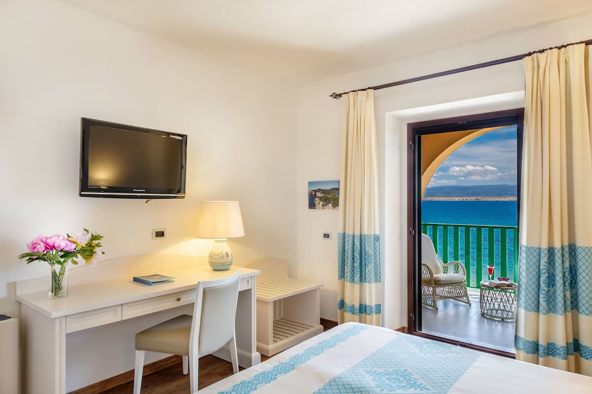 Ein Hotelzimmer mit Bett, Schreibtisch mit Stuhl, Fernseher und einem Balkon mit Meerblick.