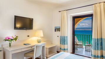 A hotel room with a bed, a desk, a chair, a television, and a balcony with a view of the sea.