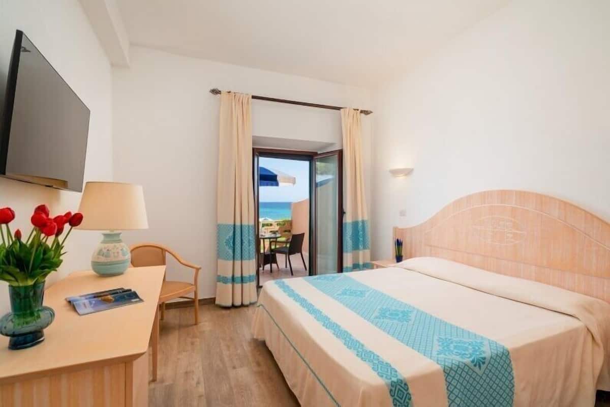 Ein Hotelzimmer mit Bett, Schreibtisch mit Lampe, Stuhl, Fernseher und Meerblick durch eine Glastür.