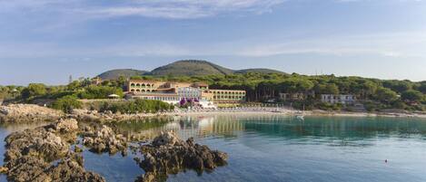 A coastal hotel with a clear view of the sea, rocky shoreline, and a hilly backdrop.