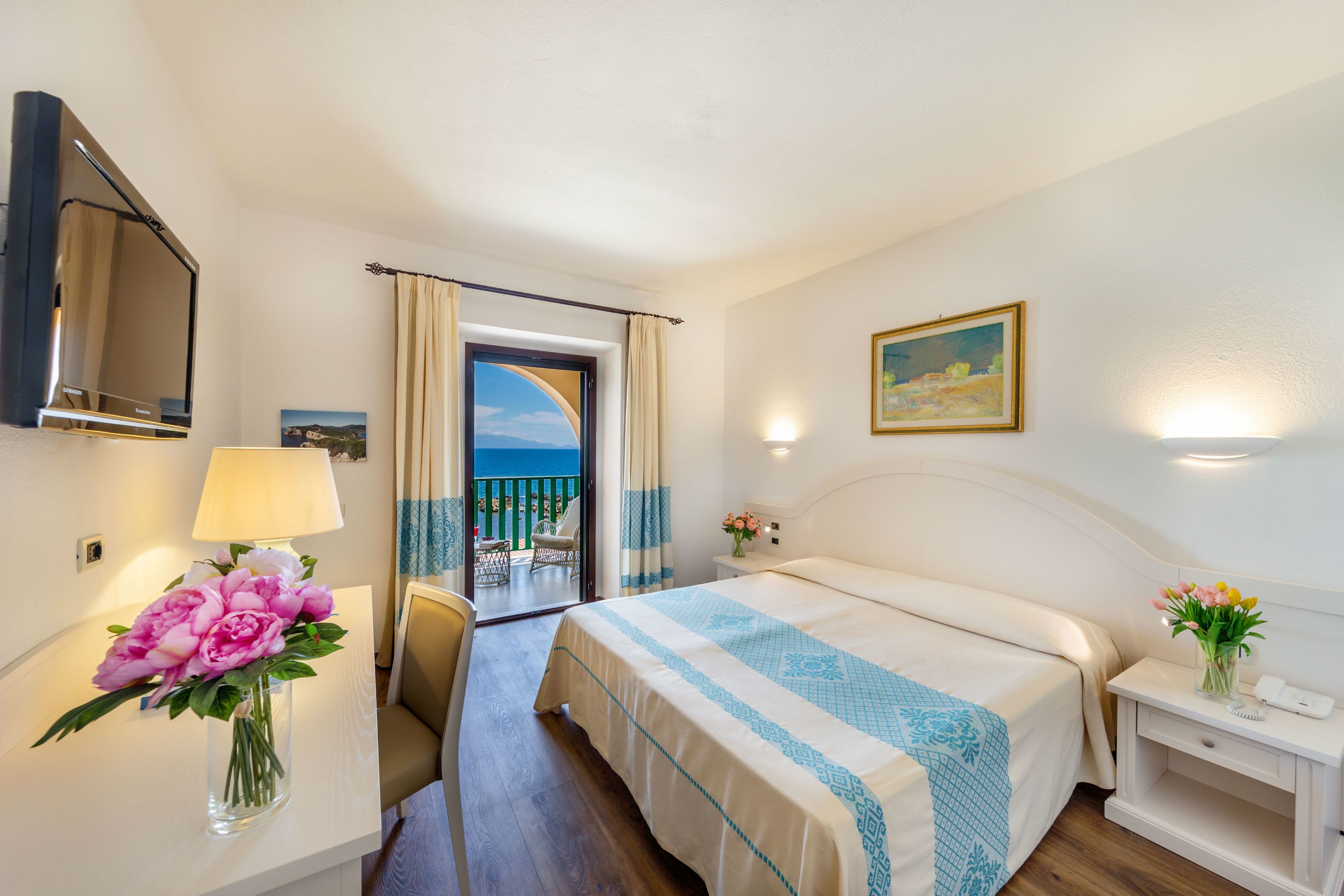 A hotel room with a bed, a desk with a lamp and flowers, a chair, a nightstand with a phone, and a painting on the wall.