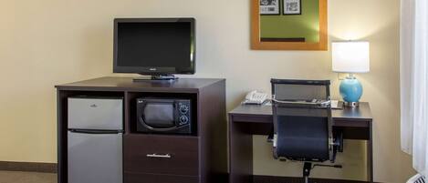 A compact hotel room with a kitchenette including a refrigerator, microwave, and a flat-screen TV. There's a desk with a chair and a lamp, and a mirror on the wall.