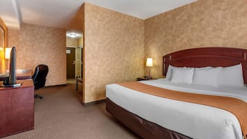 A hotel room with a large bed, a desk with a computer, a chair, and a nightstand with a lamp.