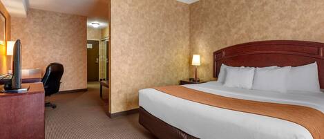 A hotel room with a large bed, a desk with a computer, a chair, and a nightstand with a lamp.