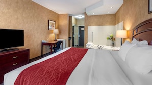 A hotel room with a large bed, a desk with a chair, a television, a bathroom with a mirror, and a door leading to another room.