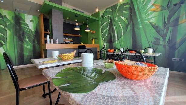Un comedor con una mesa dispuesta para dos personas, con un centro de mesa de hoja verde grande, una jarra blanca y cestas anaranjadas.