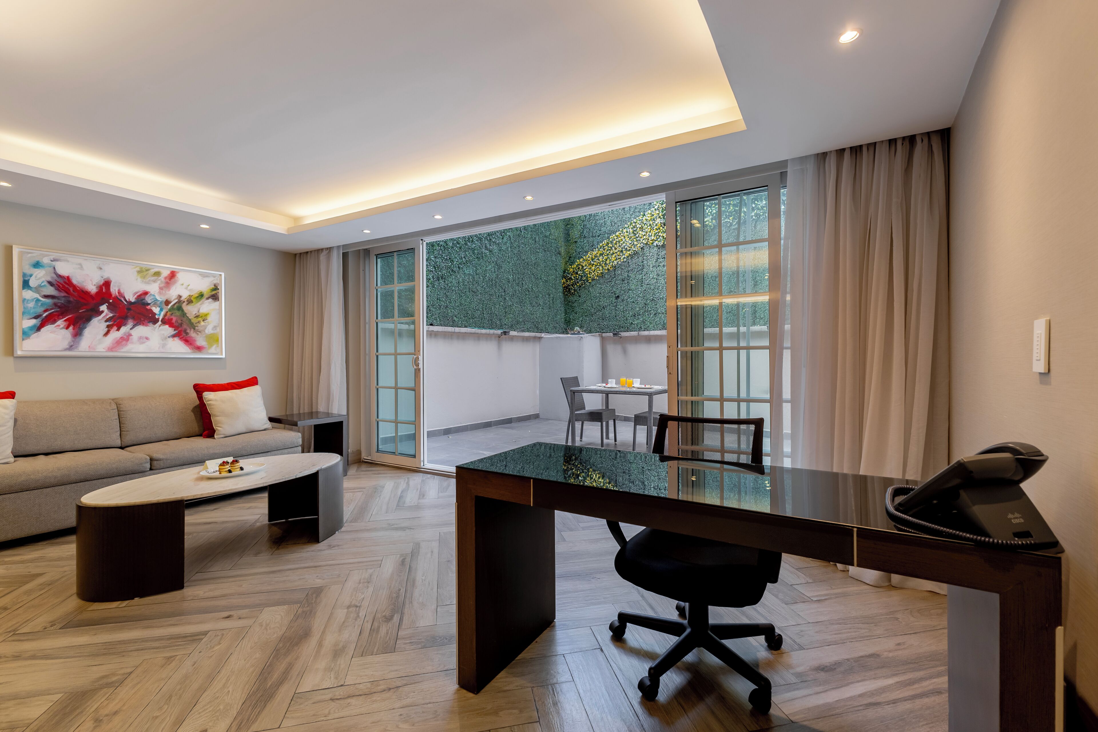 A modern hotel room with a sofa, a glass-top desk, a dining area, and a large window with curtains.