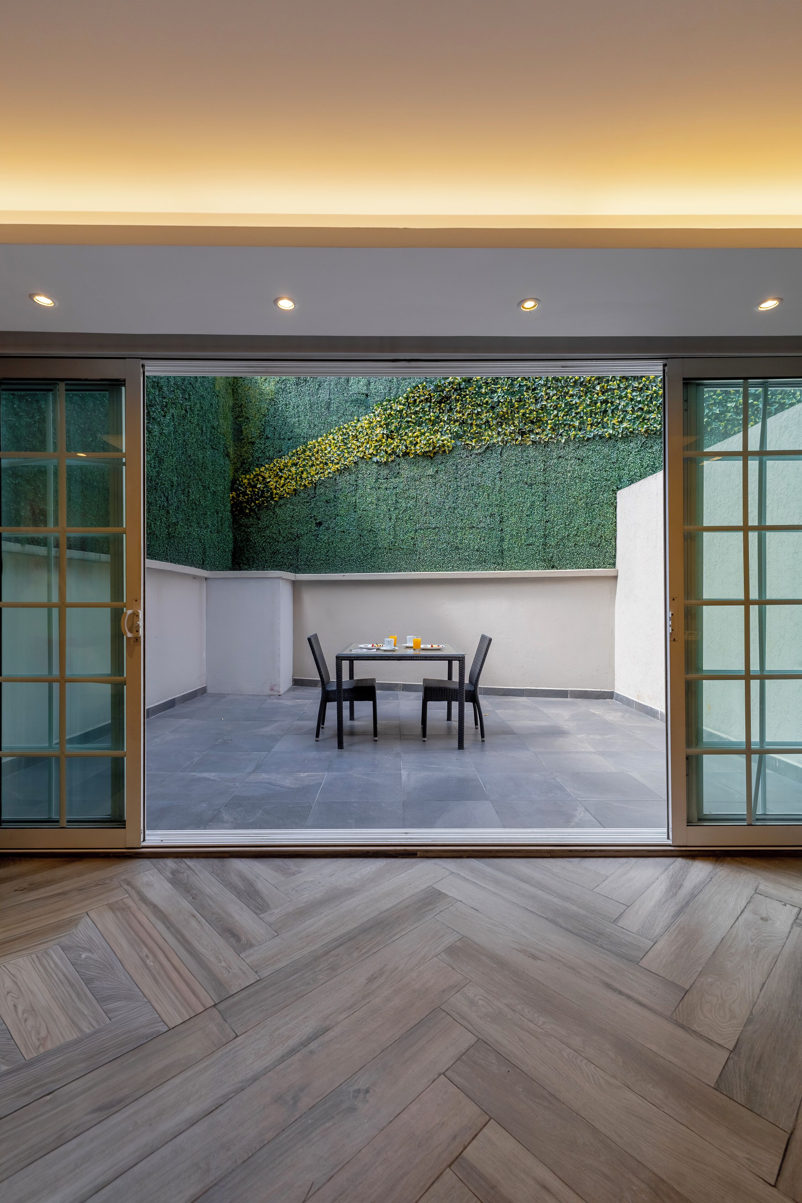 A modern patio with a table and chairs, a green wall with yellow flowers, and a herringbone wood floor.