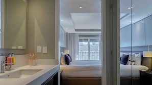 Una habitación de hotel moderna con un espejo grande, piso de madera en patrón espiga, cama con vista al balcón y baño independiente con tocador y lavamanos.