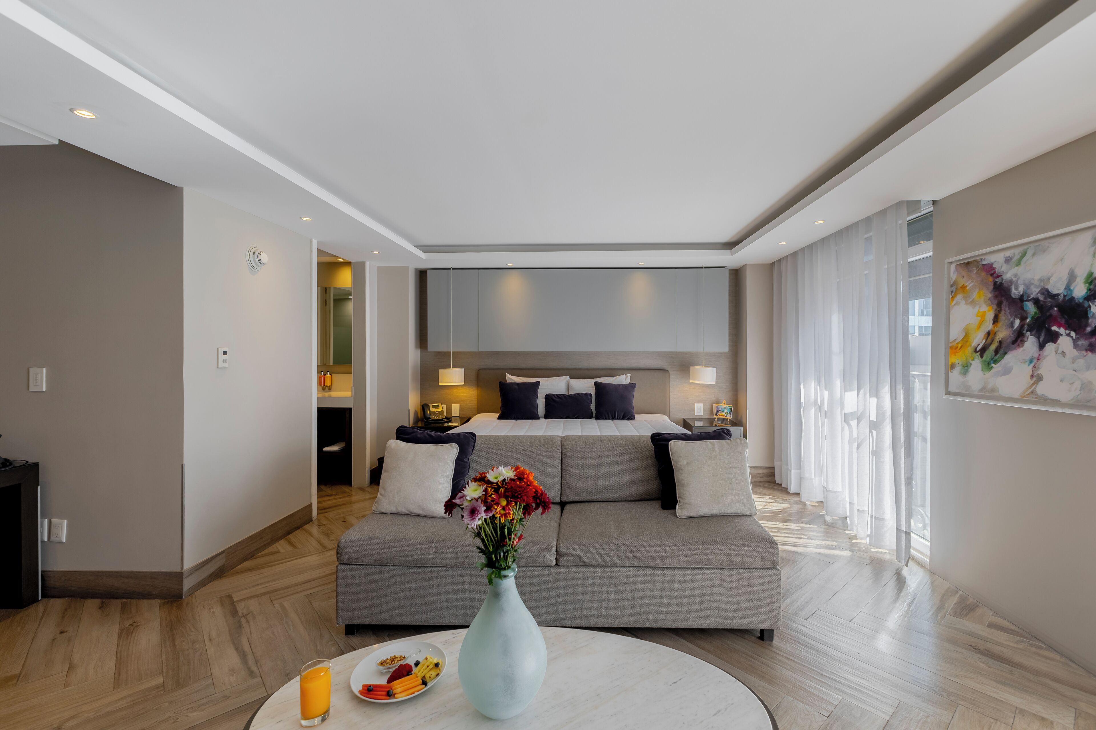 Una habitación de hotel moderna con un sofá gris, una mesa de comedor con fruta, un jarrón de flores y una cama con cabecera.