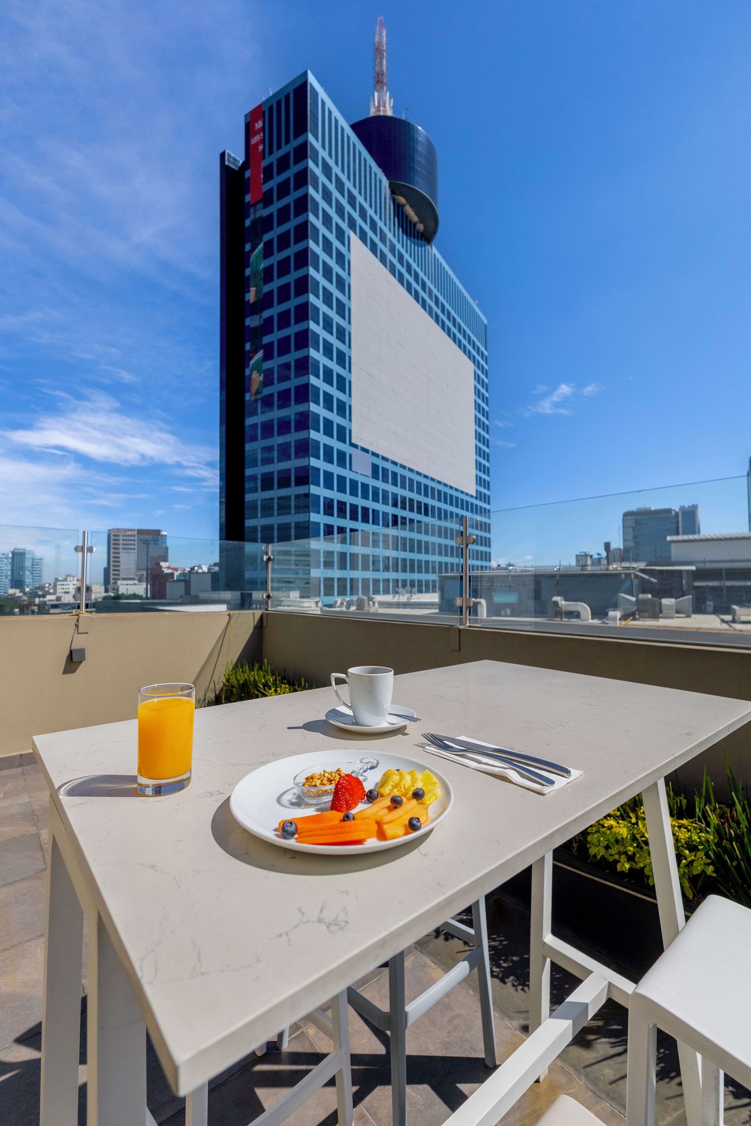 Une terrasse sur le toit, avec une table dressée pour le petit-déjeuner, un verre de jus d’orange et une assiette de fruits. On aperçoit, en arrière-plan, un bâtiment moderne à la façade bleue et un grand panneau publicitaire.