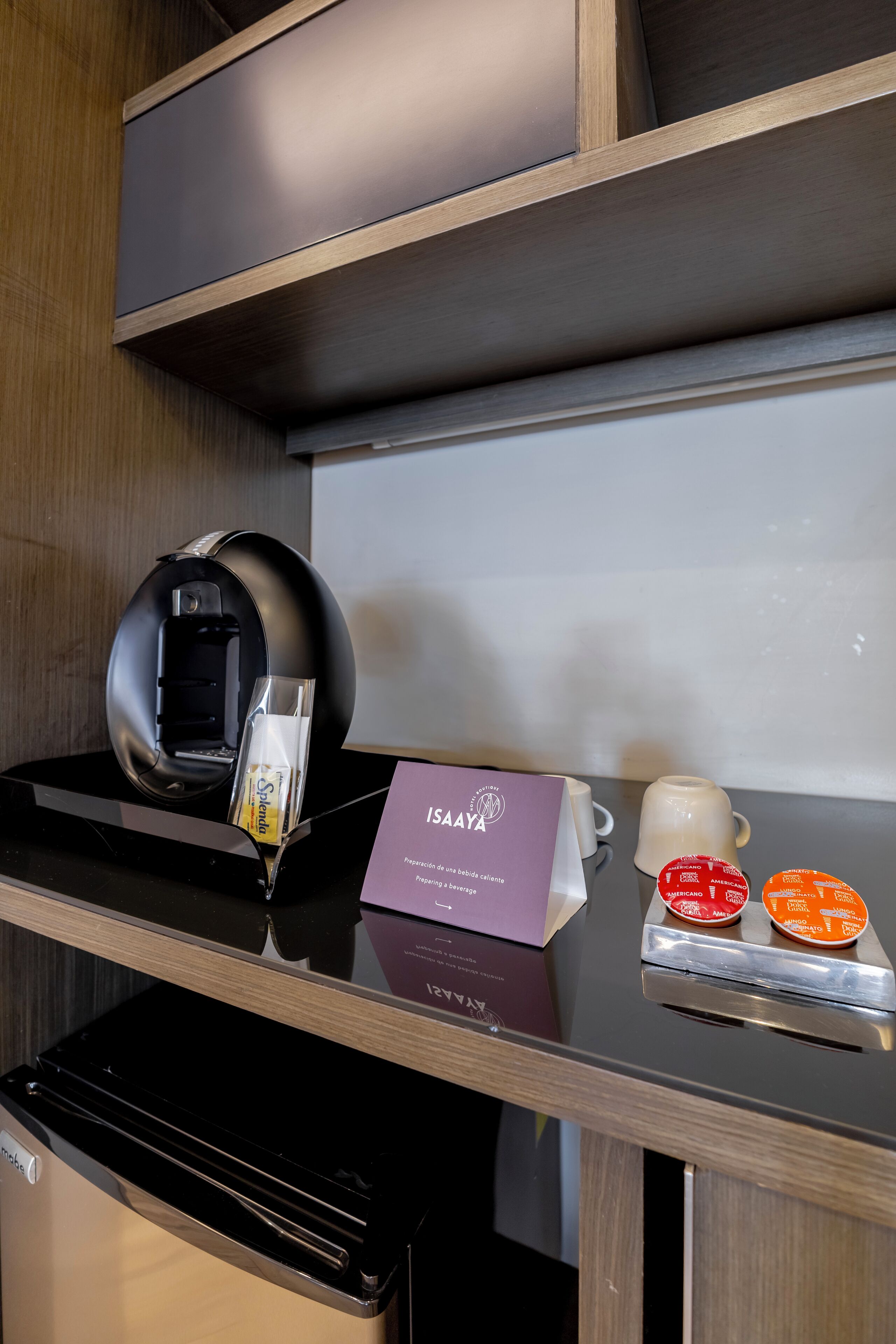 Un minibar con una cafetera, un vaso de agua, una taza de café y un cartel de ISAAYA.