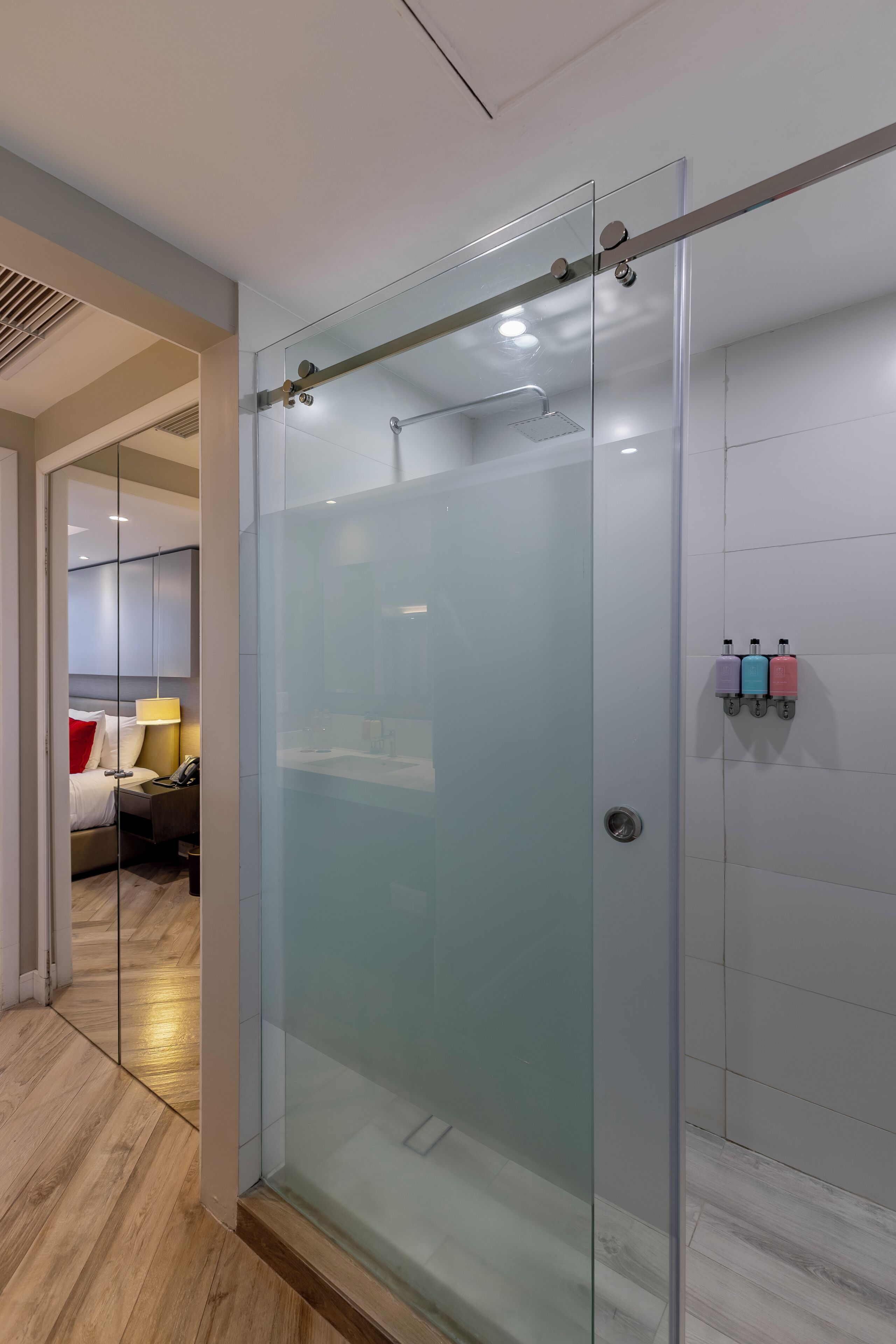 A modern hotel room with a glass-enclosed shower, a bed with a red pillow, and a bedside table with a lamp and phone.