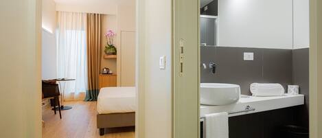 A hotel room with a bed, a desk, a chair, a bathroom with a sink, mirror, and towel rack.