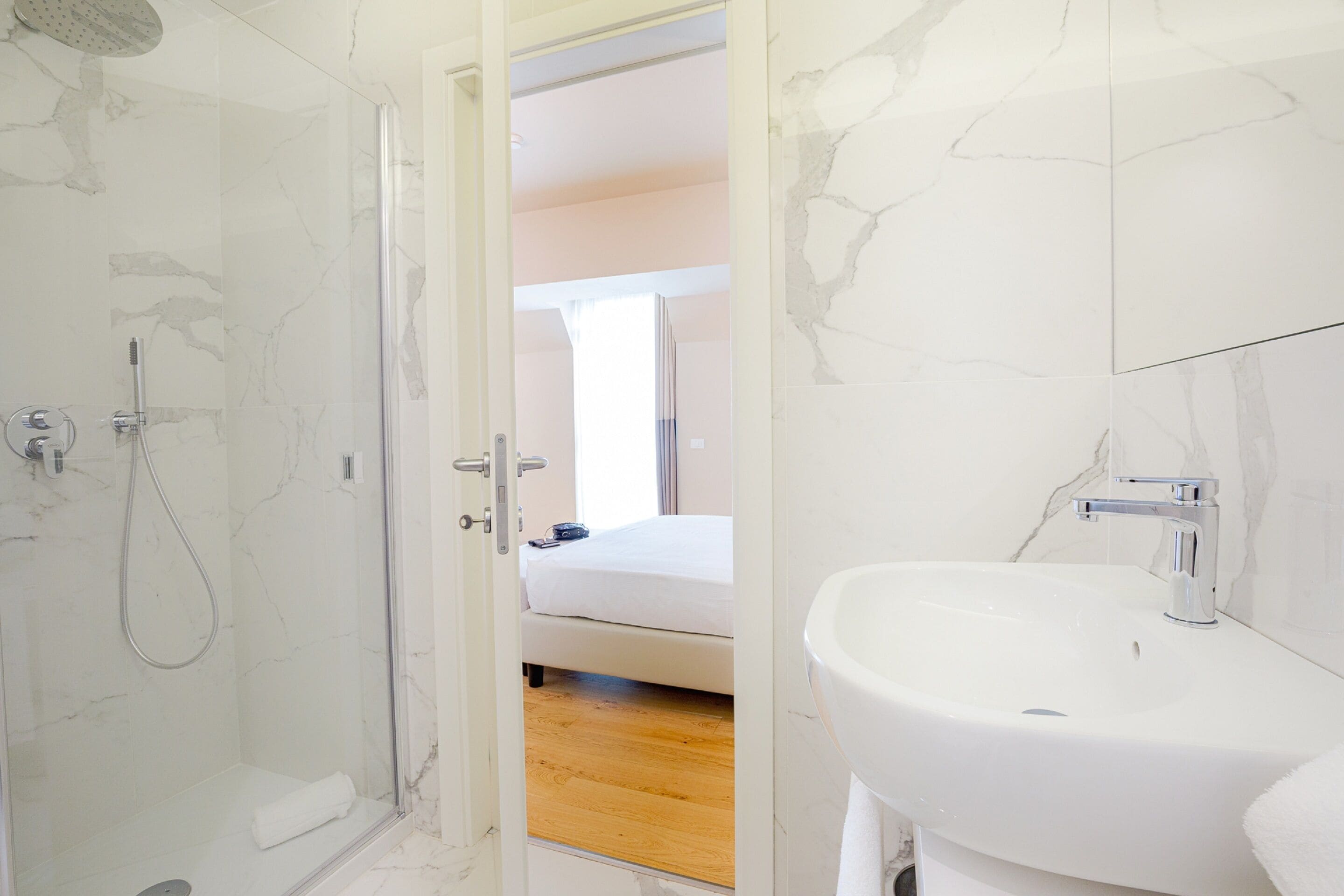 Un bagno con doccia vetrata, un lavandino bianco e un letto visibile attraverso una porta aperta.