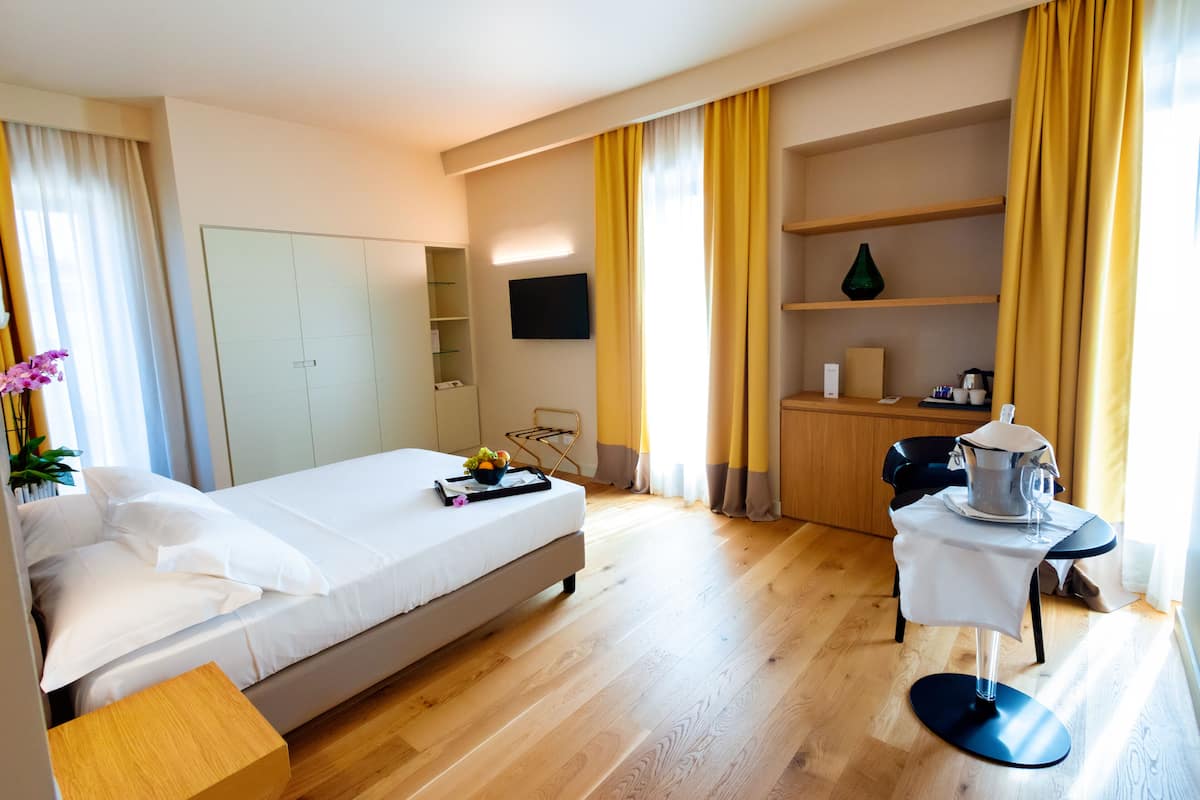 Una habitación de hotel con cama, escritorio, silla, televisor y repisa con un florero.