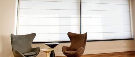 A room with two modern chairs and a small table, large windows with blinds, and wooden flooring.
