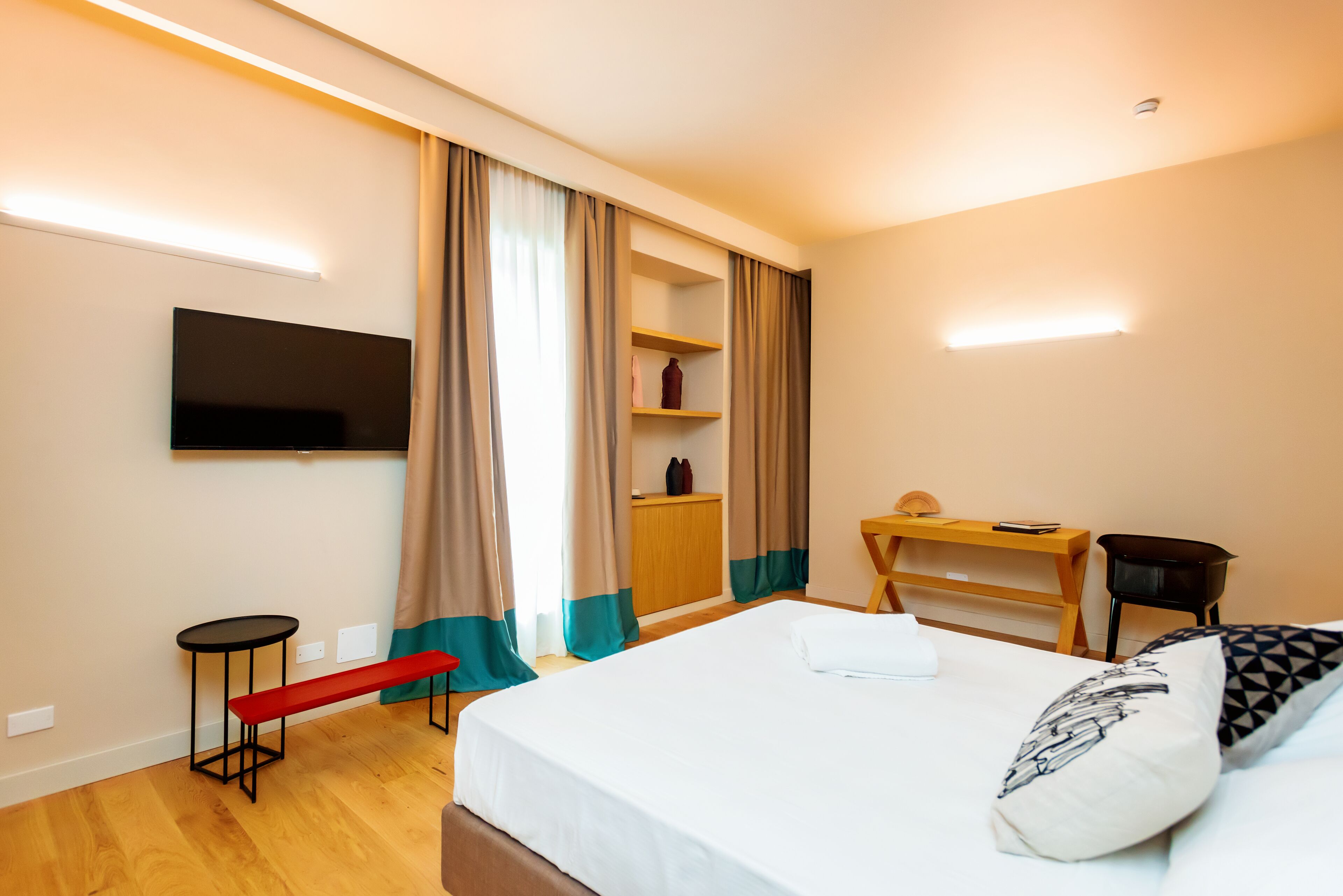 A hotel room with a bed, a TV mounted on the wall, a small desk, a bench, and a wardrobe.