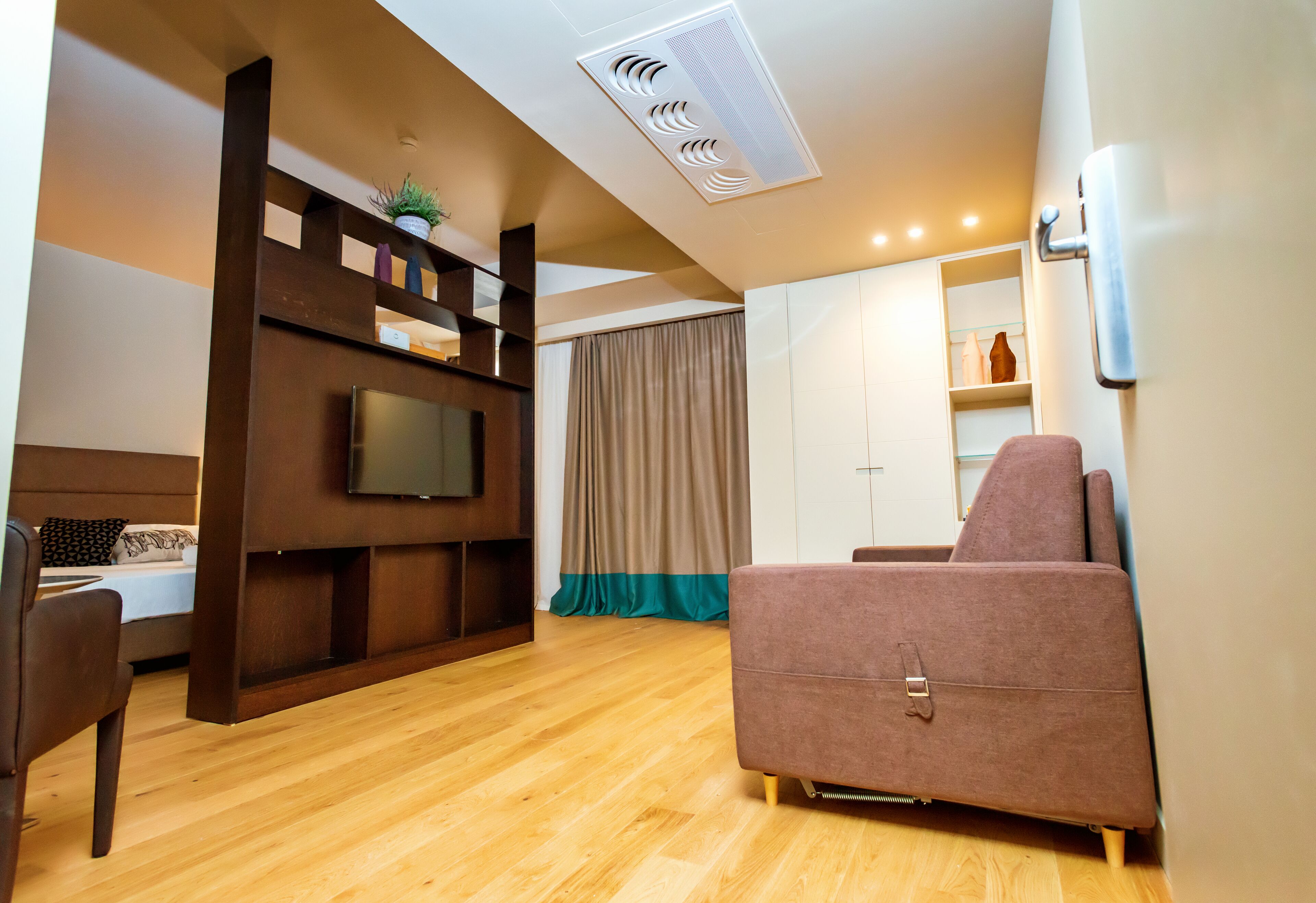 Ein modernes Wohnzimmer mit Holzboden, einem wandmontierten Flachbildfernseher, einem braunen Sofa, einem Esstisch mit Stühlen und einem Kleiderschrank mit Spiegel.