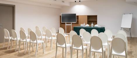 A conference room with white chairs arranged in rows, a table with a green tablecloth, a large flat-screen TV, and a whiteboard.
