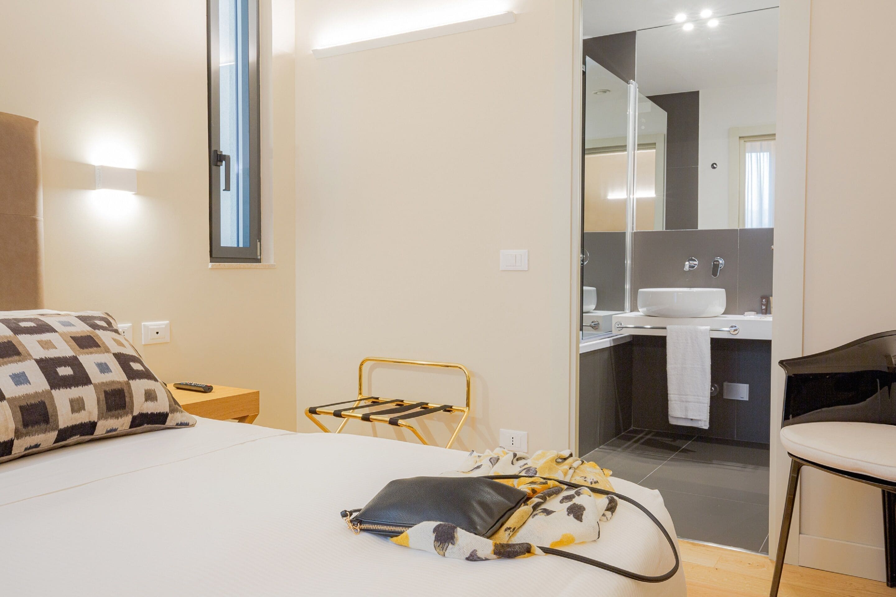 A hotel room with a bed, a bedside table, a chair, a bathroom with a sink and mirror, and a towel hanging on the wall.