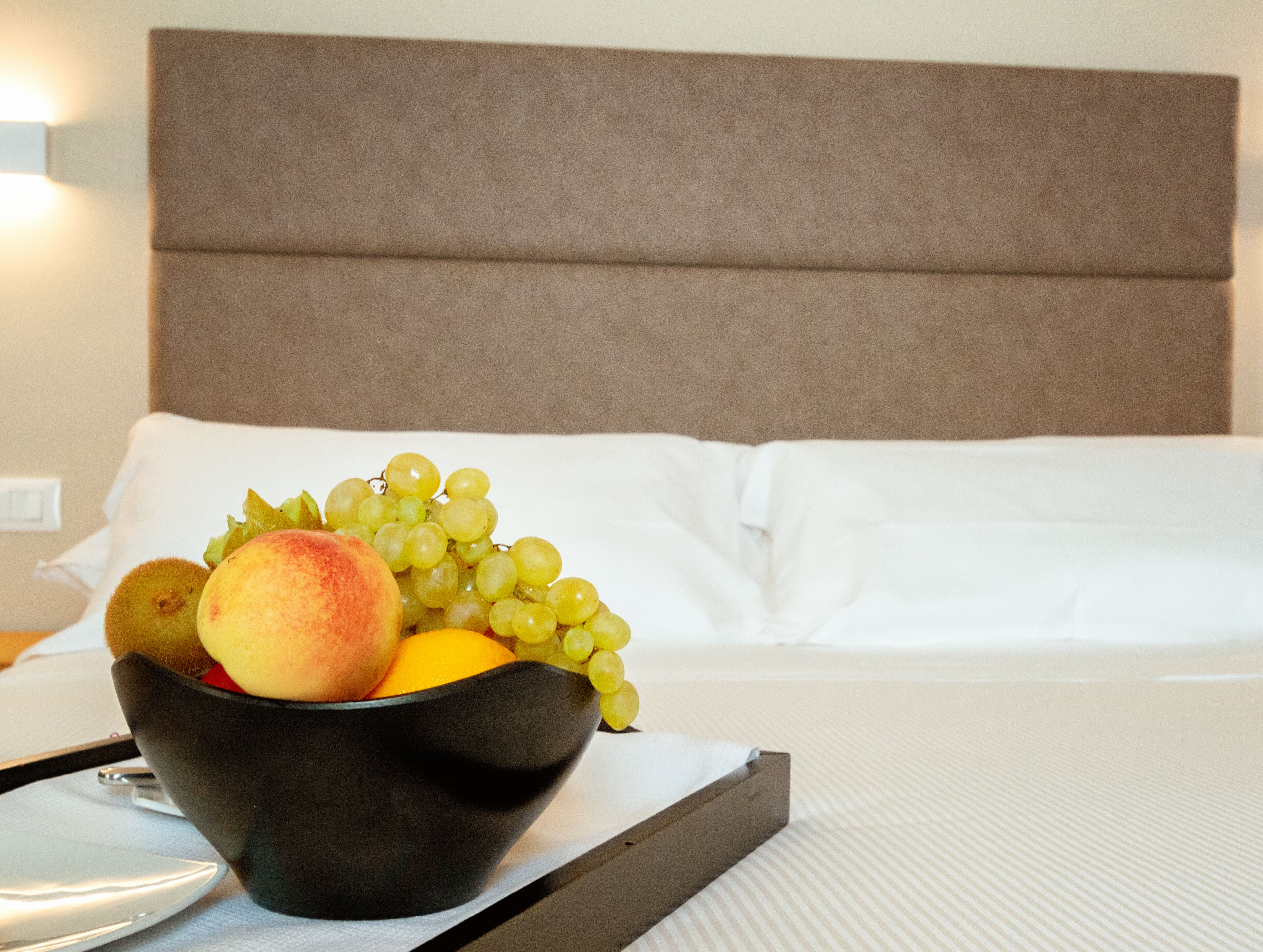 Un lit avec du linge de lit blanc et une tête de lit, un plateau avec un bol de fruits et une table de chevet avec une lampe.