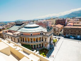 Un edificio storico con una cupola e architettura classica, immerso tra strade cittadine con montagne sullo sfondo.