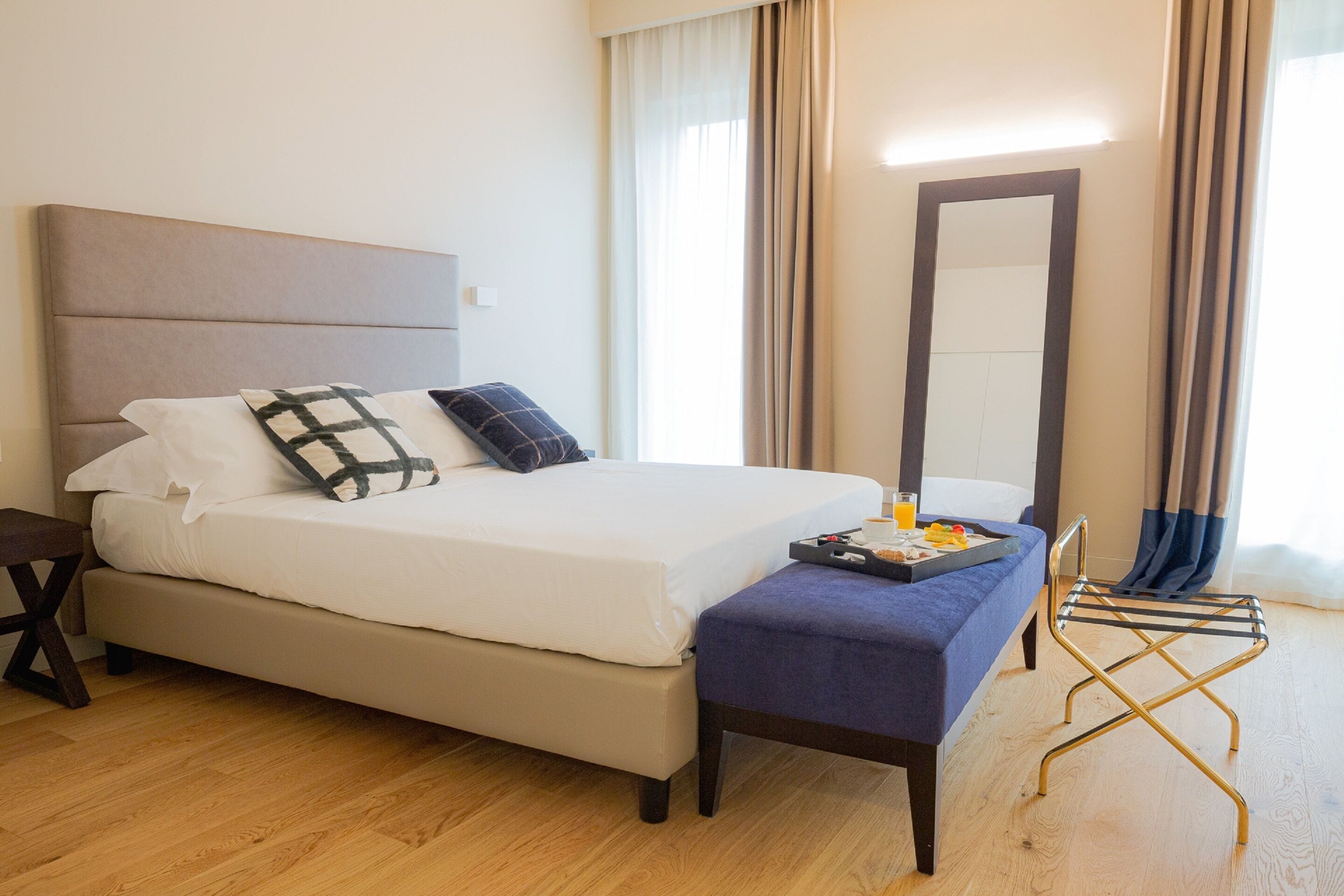 A neatly made bed with white linens and two pillows, a bedside table with a tray of breakfast items, a bench with a tray, and a mirror on the wall.