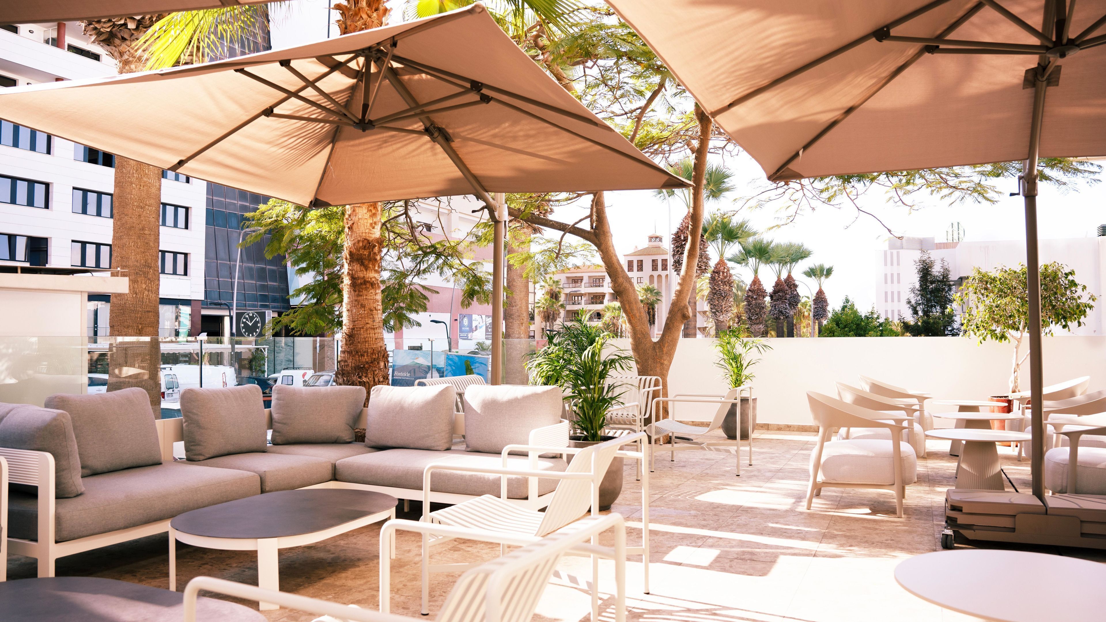 En tagterrasse med udendørs møbler, herunder sofaer og borde, overdækket af store parasoller, omgivet af palmer og bygninger.