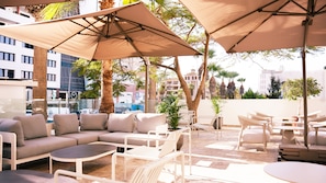 Terraza en la azotea con mobiliario exterior, incluyendo sofás y mesas, protegida por grandes sombrillas y rodeada de palmeras y edificios.